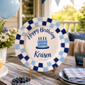A personalized birthday plate with a blue and white checkered border, displayed on a decorated outdoor porch table. The plate features a blue and white checkered border, a central illustration of a birthday cake with candles, and the text 'Happy Birthday Kaisen' along with the date '07.