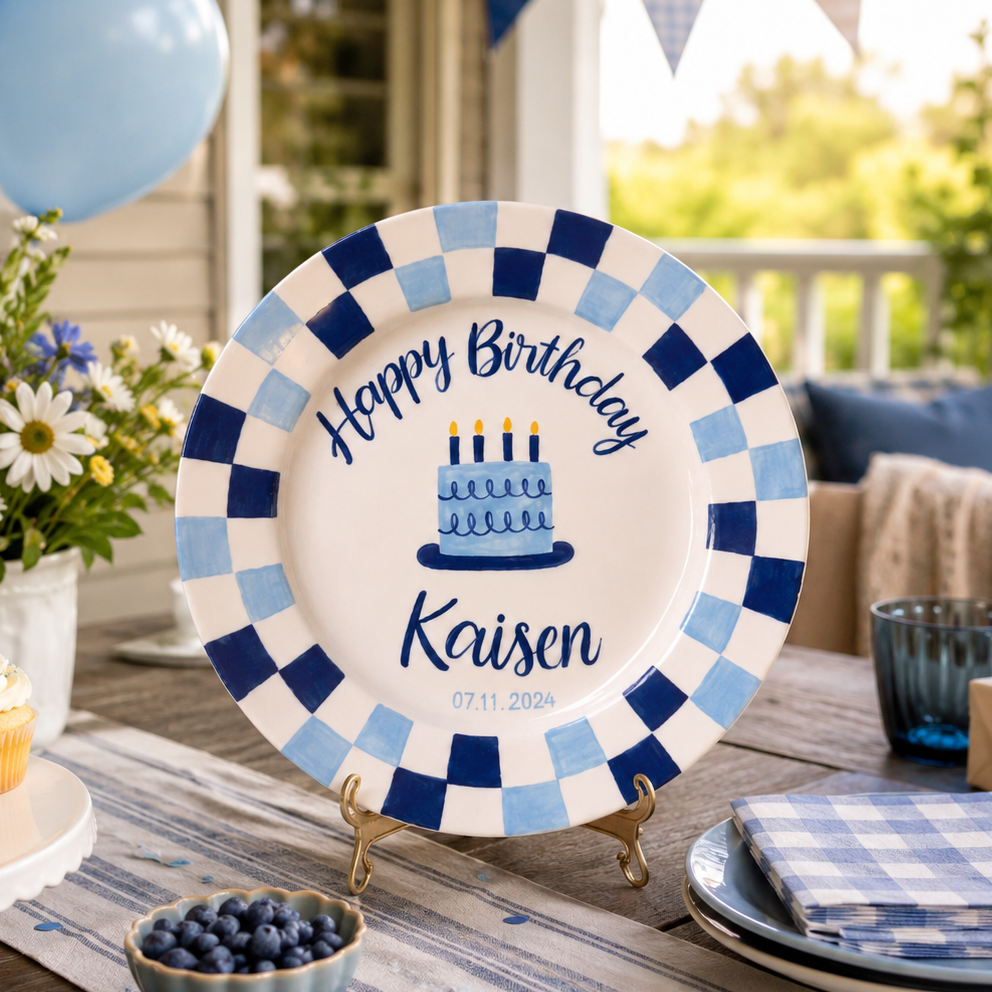 A personalized birthday plate with a blue and white checkered border, displayed on a decorated outdoor porch table. The plate features a blue and white checkered border, a central illustration of a birthday cake with candles, and the text 'Happy Birthday Kaisen' along with the date '07.