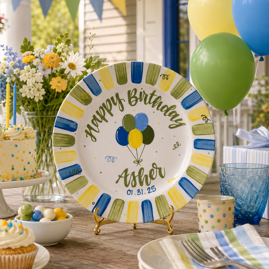 A round white birthday plate on a small gold stand, printed with 'Happy Birthday Asher' and a balloon graphic in blue, yellow, and green, displayed on a decorated outdoor porch table with cupcakes, balloons, and a cake.